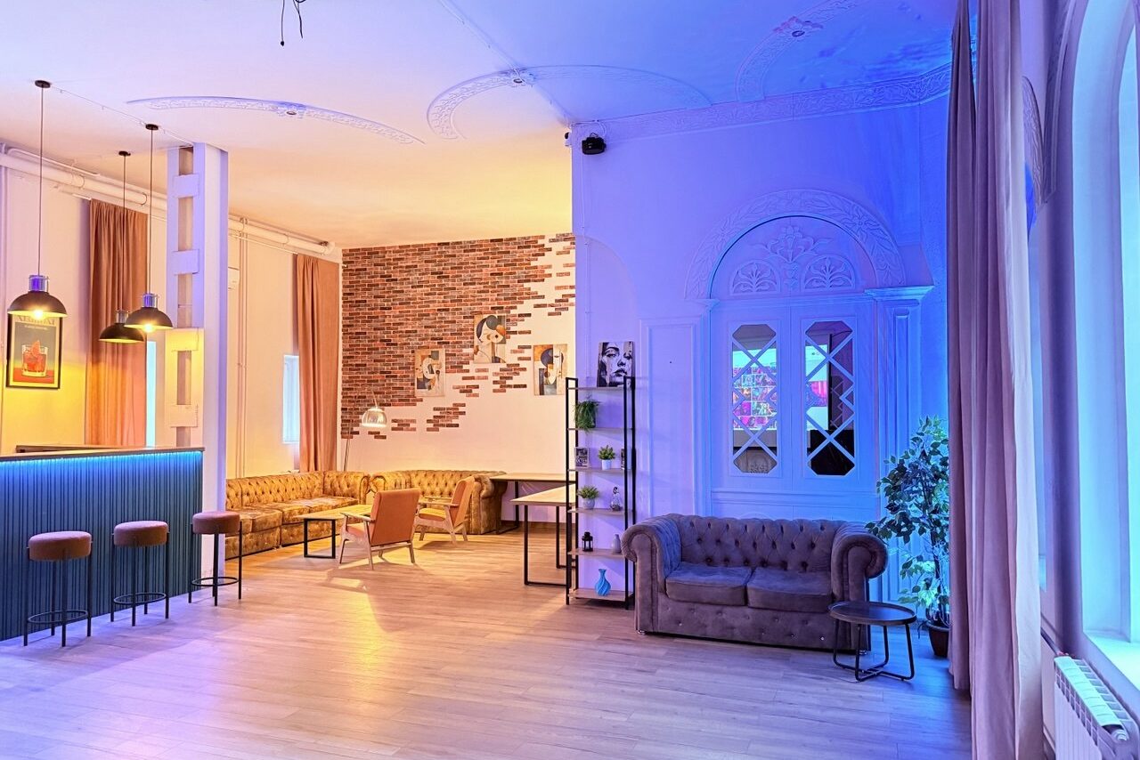 Арендовать Лофт Софт. Лофт-пространство с барной стойкой, барными стульями, диванами, столиками и стеллажом. Открытая планировка, декоративные кирпичные стены, лепной потолок. Освещение разноцветное, теплое и неоновое. Паркетный пол, окна с занавесками, комнатные растения, постеры.