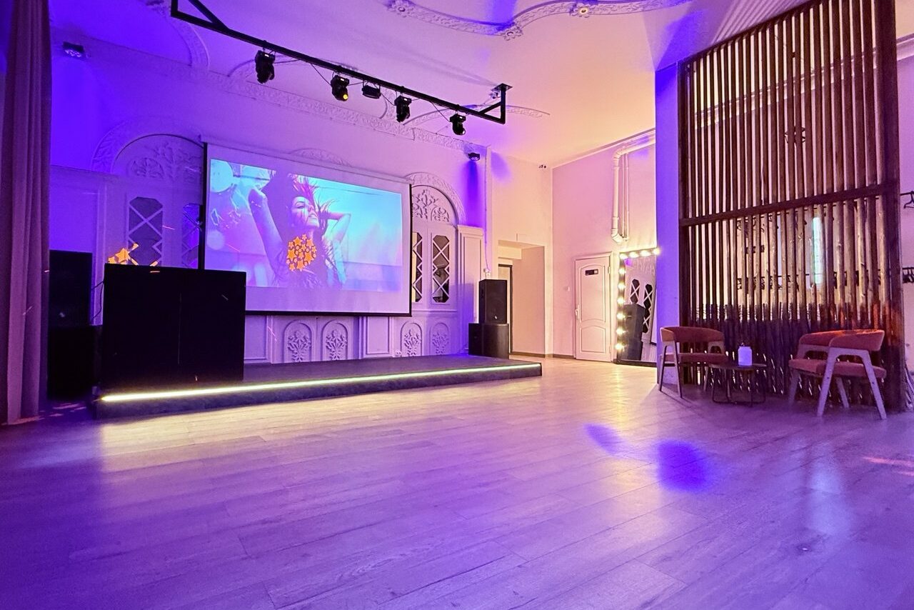 Арендовать Лофт Софт. Просторное помещение с деревянным полом, сценой и большим экраном для проекций. Освещение разноцветное, фиолетовые и теплые тона. Справа несколько стульев, на стене декоративные деревянные панели. В комнате потолочные светильники.