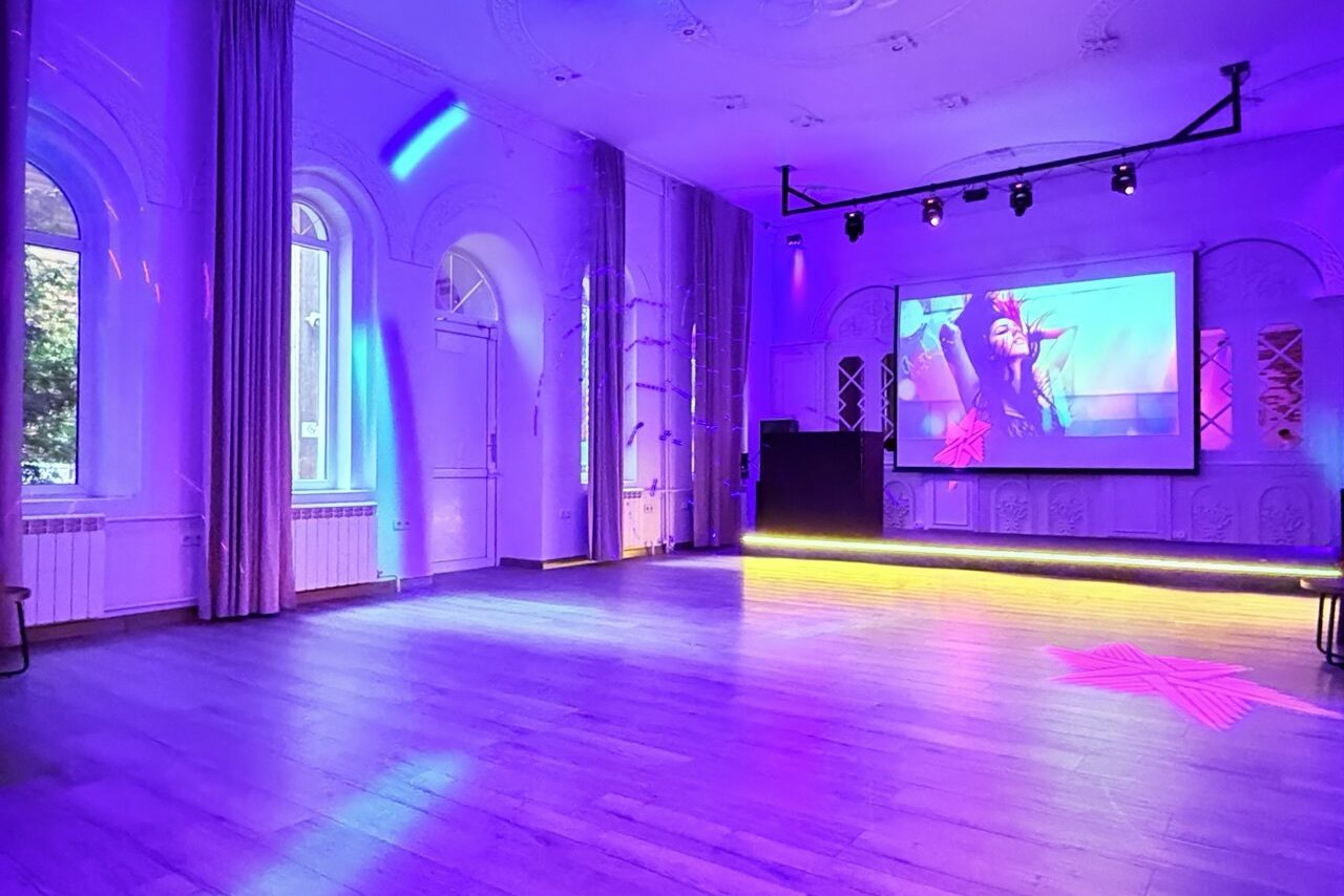 Арендовать Лофт Софт. Просторный зал с деревянным полом, высокими потолками и большими арочными окнами. На стене висит крупный экран с изображением. В помещении фиолетовая и синяя подсветка, световые приборы под потолком. Стены белые, украшенные рельефами.