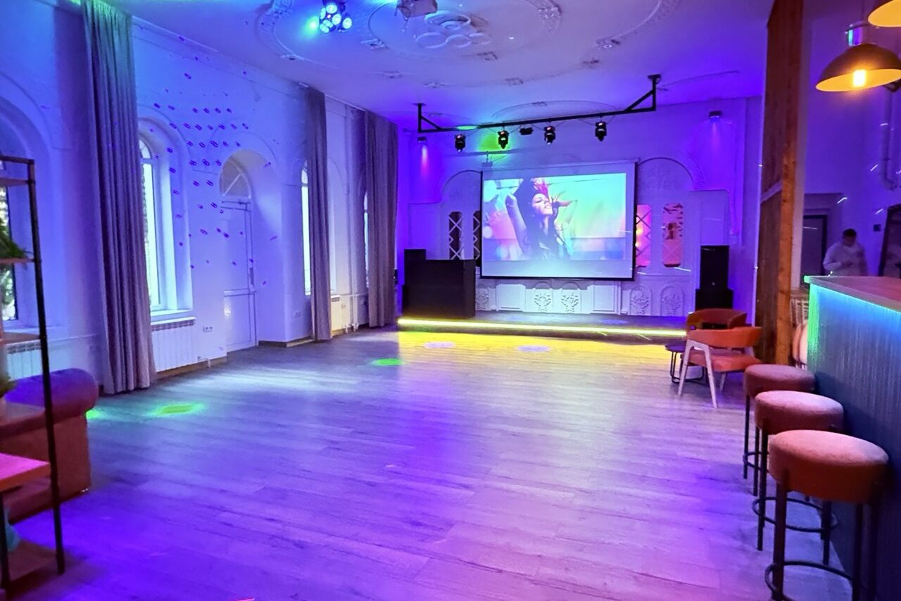 Арендовать Лофт Софт. Просторное помещение с деревянным полом, центральной колонной, барной стойкой и стульями. На стене большой экран с проектором. Разноцветное освещение, окна с шторами. Потолок украшен лепниной. Мягкие стулья и диван у стены.