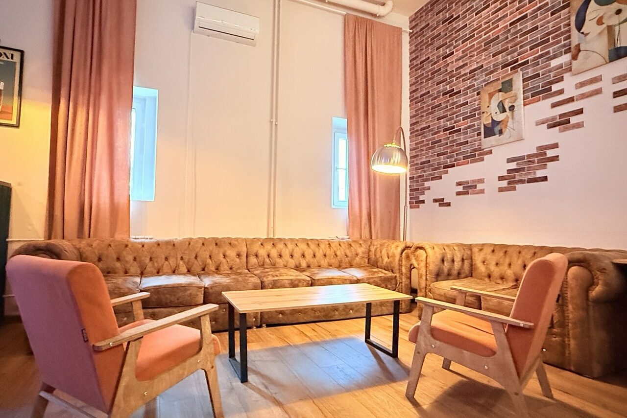 Арендовать Лофт Софт. Уютное пространство с мягким диваном угловой формы и двумя креслами с деревянными подлокотниками. Стены частично отделаны кирпичом, светлые занавески, висячая лампа. На деревянном полу ковёр. На стене картина и окно с белыми рамами.