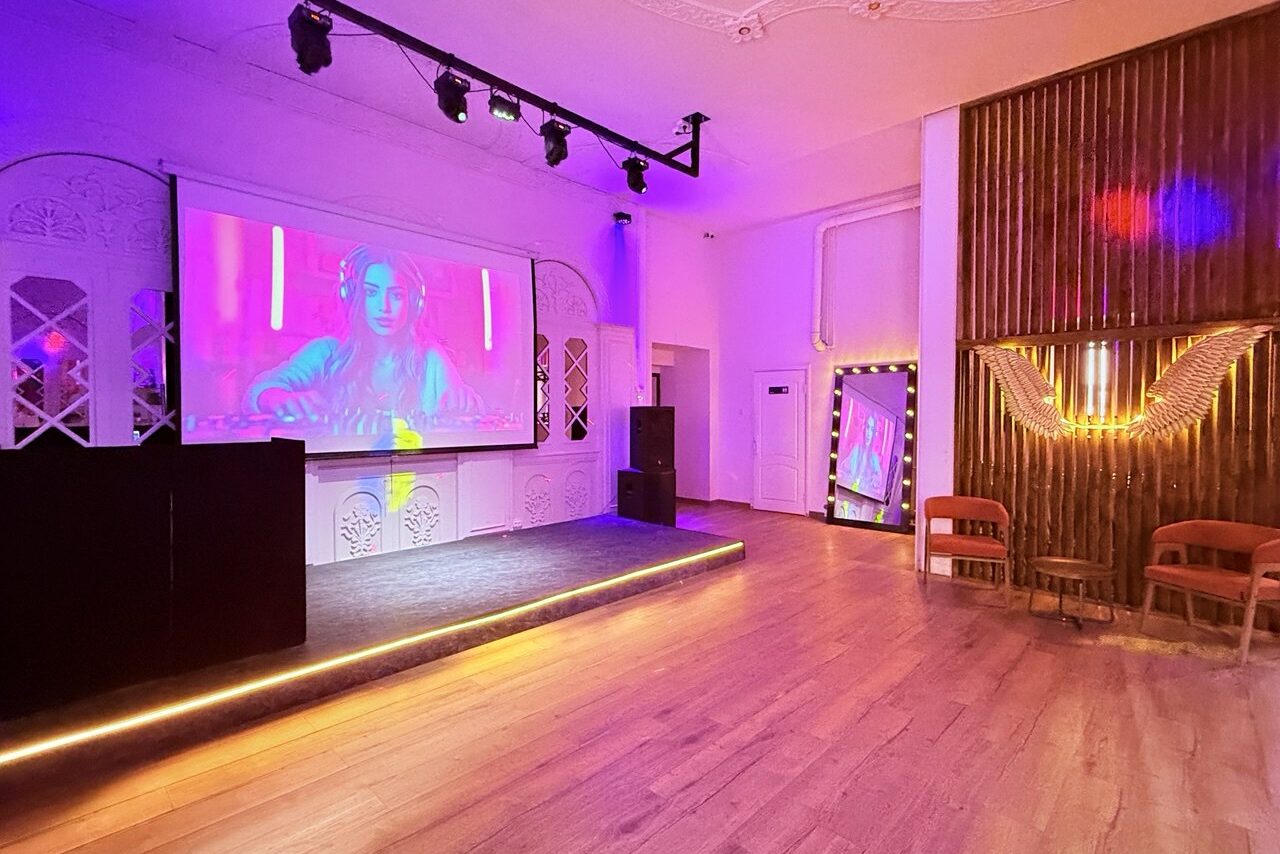 Арендовать Лофт Софт. Пространство с деревянным полом и сценой, на стене большой экран с проекцией. Слева черное оборудование, справа зона с двумя стульями и большим зеркалом в подсветке. Потолочные светильники и разноцветное освещение.