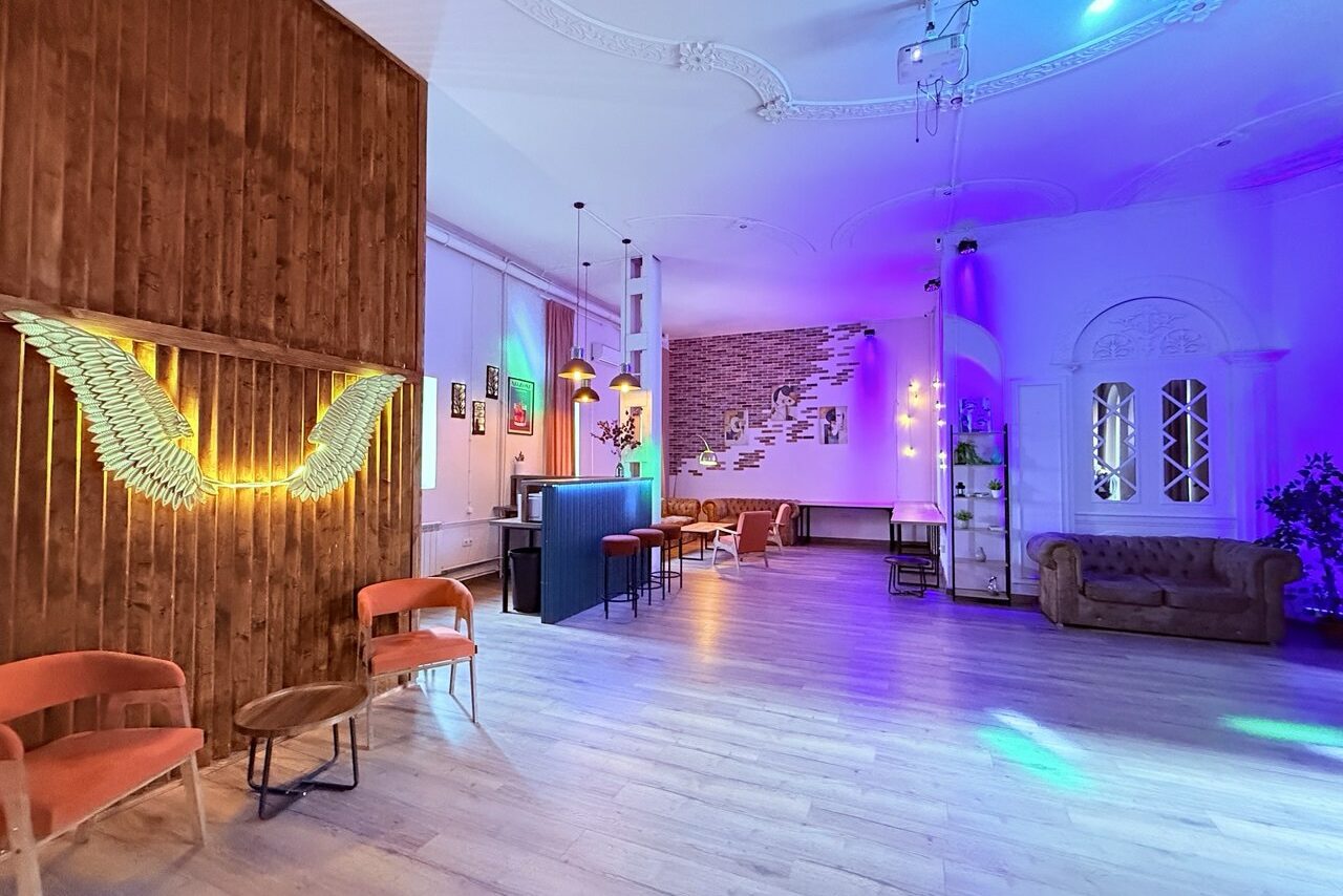 Арендовать Лофт Софт. Просторное помещение с деревянными и кирпичными стенами, разноцветная подсветка. Столы и стулья, мягкие кресла и диван. Потолочный декор, картина на стене и неоновый светильник в форме крыльев. Светлый ламинат на полу.