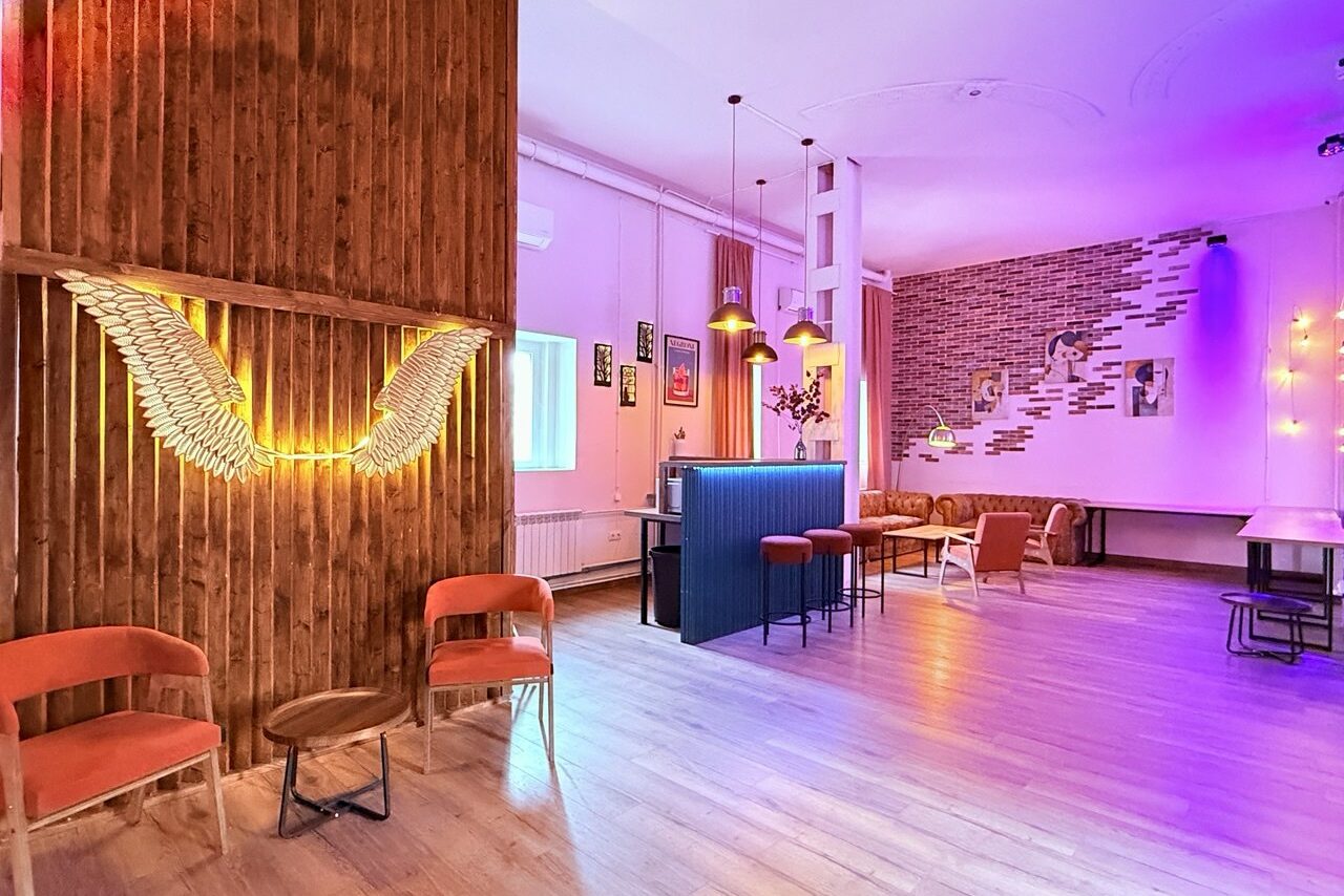 Арендовать Лофт Софт. Просторная комната с деревянной и кирпичной отделкой, декор в форме крыльев на стене, разноуровневое освещение с яркими цветами. Мебель: барная стойка, стулья, диваны. Стены украшены картинами, светодиодные гирлянды. Пол паркетный.