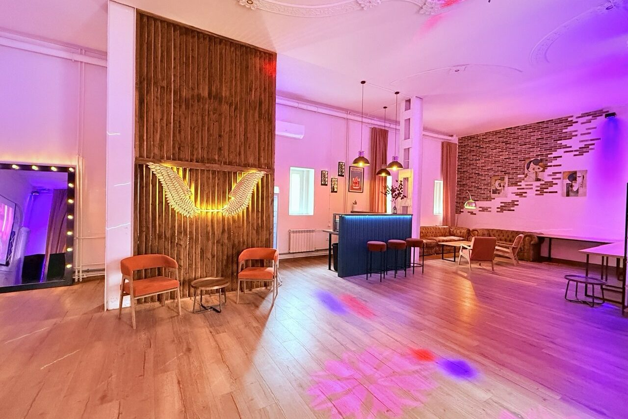 Арендовать Лофт Софт. Многофункциональное помещение с деревянным полом, зонами отдыха и барной стойкой. Яркое неоновое освещение, подвесные светильники, кирпичная и деревянная отделка стен. Разнообразная мебель: стулья, диван, столы. На стене светящаяся инсталляция.