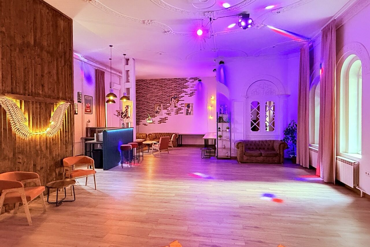 Арендовать Лофт Софт. Просторное помещение с барной зоной и лаунж-зоной. Светлая плитка на полу, мягкие кресла и диван. Деревянные панели на стенах, неоновые и разноцветные светильники. Арочные окна с занавесками, кирпичная декоративная стена.