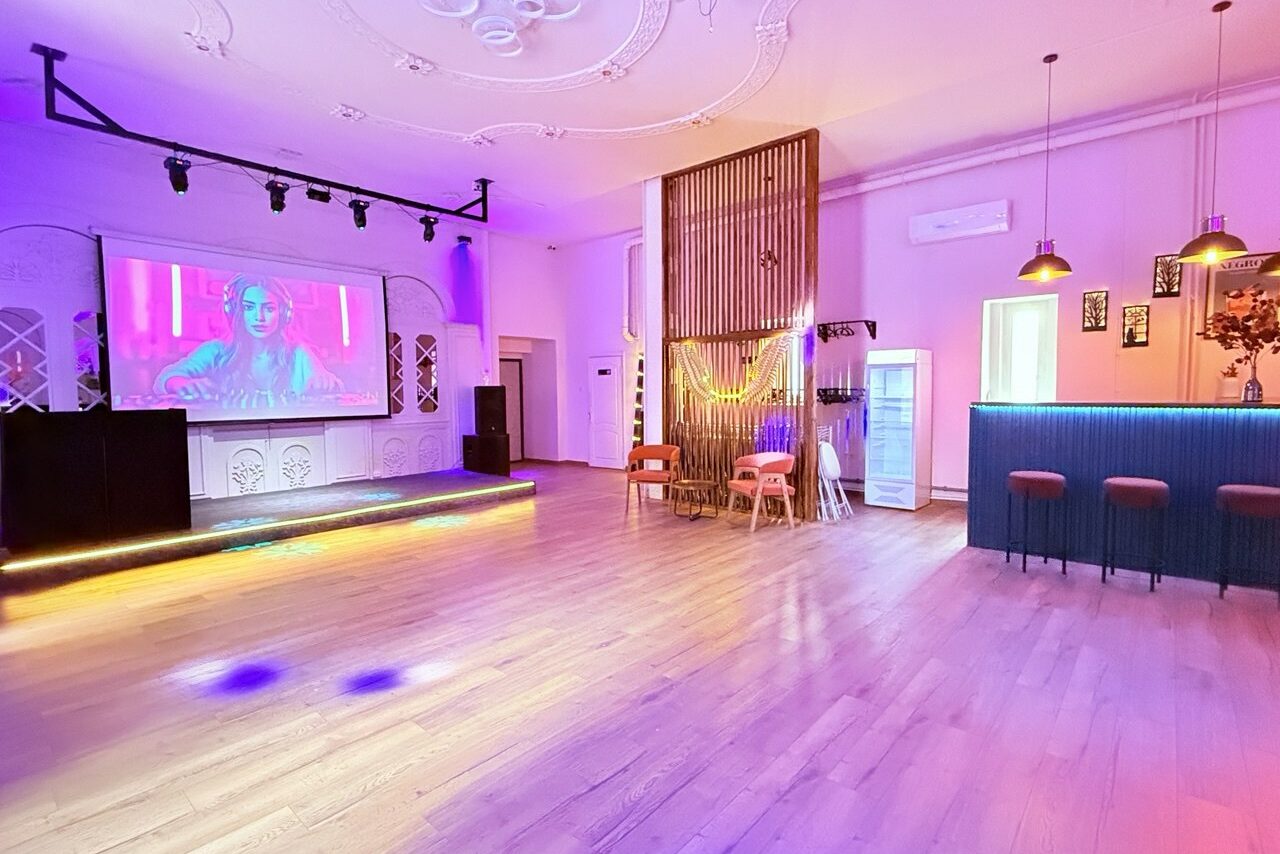 Арендовать Лофт Софт. Просторное помещение с деревянным полом, сценой с экраном, барной стойкой с высокими стульями, разноцветное освещение, декоративные панели на стене, подвесные светильники, холодильник, розовые и фиолетовые оттенки, кондиционер, несколько стульев.