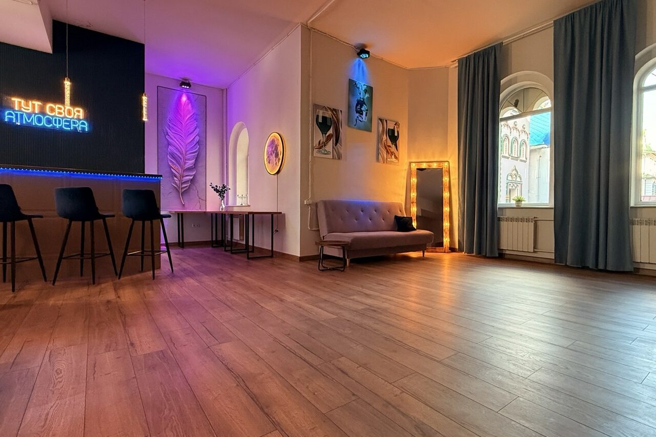 Арендовать Лофт Лайф. Помещение со светлыми стенами и деревянным полом. Имеется барная стойка с тремя черными стульями, неоновая надпись, диван, зеркало с подсветкой, стол, декор. Окна с арками и шторами. Разноцветное освещение.