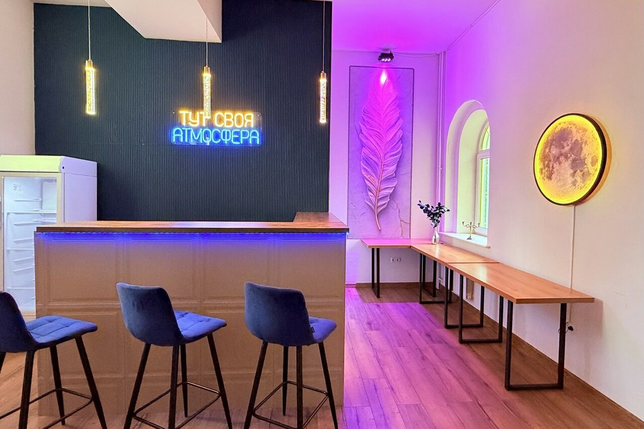 Арендовать Лофт Лайф. Барная зона с высокими стульями, неоновая надпись на тёмной стене, круглое настенное свечение, длинные деревянные столы вдоль стены, декоративные элементы, подвесные светильники, арочное окно, деревянный пол.