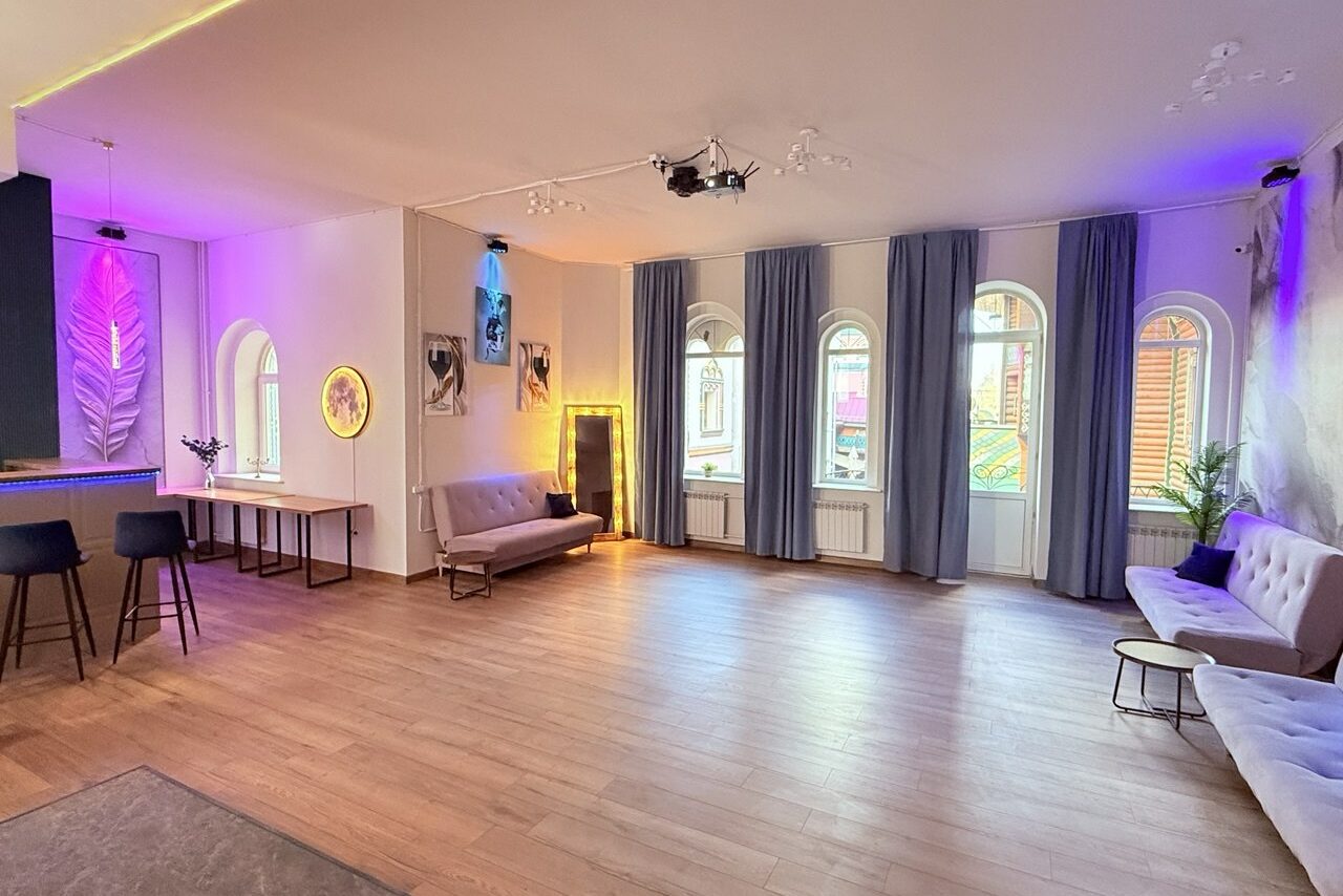 Арендовать Лофт Лайф. Просторная комната с деревянным полом и офисной мебелью. Светло-серые диваны, барная стойка с высокими стульями, неоновые огни на стенах, белые арочные окна с голубыми занавесками. Стены украшены картинами и зеркалом с подсветкой.