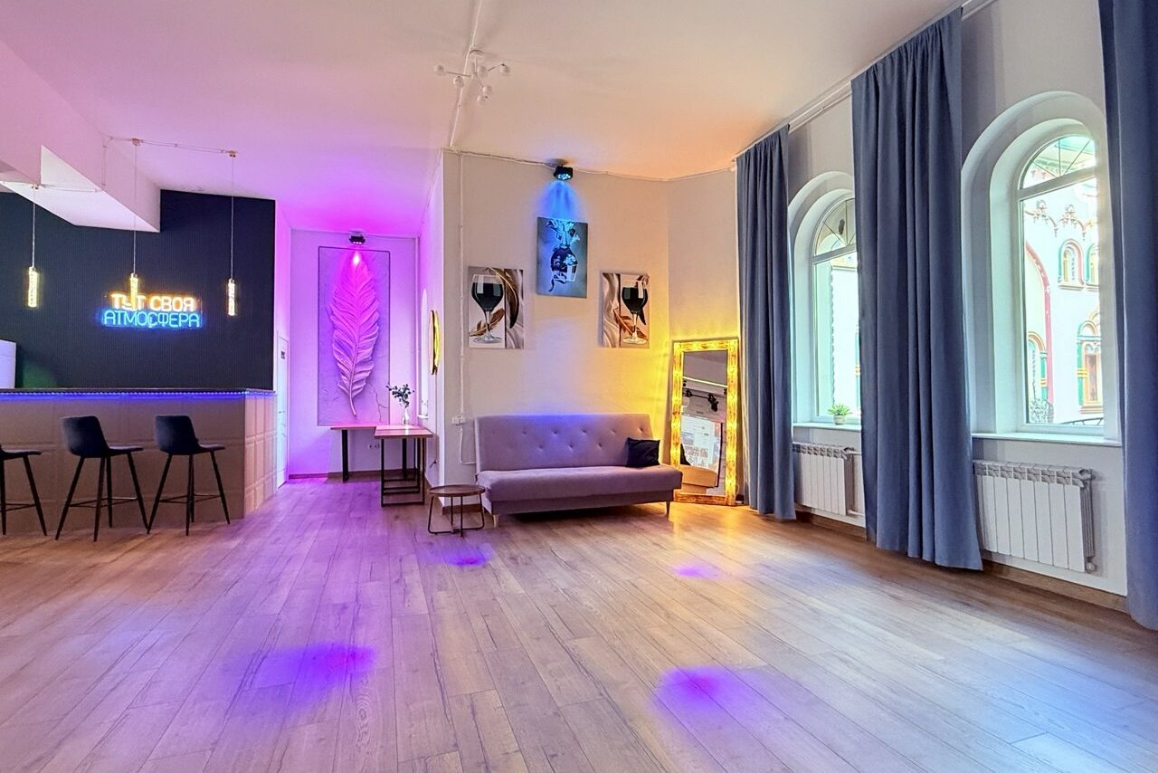 Арендовать Лофт Лайф. Просторное помещение с деревянным полом, бежевыми стенами и арочными окнами с синими занавесками. Барная стойка с двумя стульями слева. Диван, зеркало и картина на правой стене. Разноцветная подсветка и неоновая надпись на черной стене.