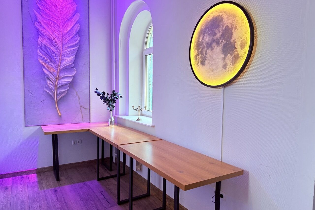 Арендовать Лофт Лайф. Комната с белыми стенами, деревянный пол. Слева деревянный стол с чёрными ножками, несколько сдвинутых столешниц. На столе ваза с цветами. На стене декоративное круглое светящееся панно и зона с перьевой декорацией.
