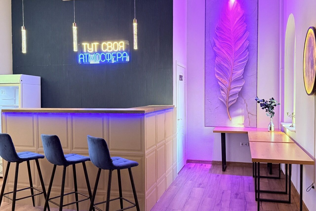 Арендовать Лофт Лайф. Барная стойка с тремя стульями на высоких ножках, неоновая вывеска на стене с надписью "Тут своя атмосфера", подвесные светильники, большая декоративная фигура в виде листа с подсветкой, деревянные столы, белые и черные стены, светлый пол.
