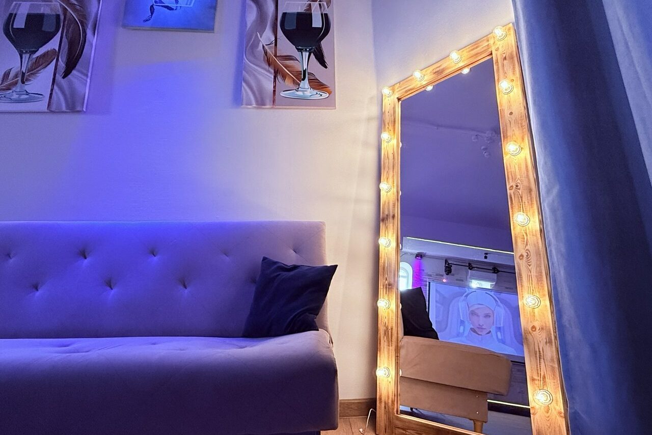 Арендовать Лофт Лайф. Комната с мягким диваном, обитым светлой тканью, темно-синим подушкой и большим зеркалом в деревянной раме с подсветкой. На стене две картины с бокалами вина. Полотно с изображением лица в отражении. Светодиодное освещение синим цветом.