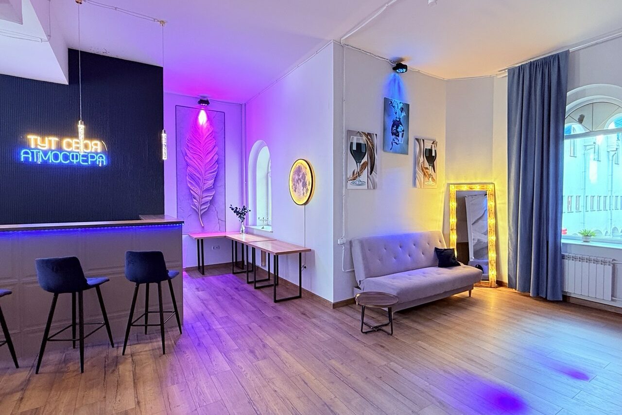 Арендовать Лофт Лайф. Современное пространство с барной зоной и трех стульями, диван, зеркалo, столы. Неоновая подсветка, окна с арками, паркетный пол. На стене постеры, надпись на стене "Тут своя атмосфера". Занавески слева, радиатор под окном.
