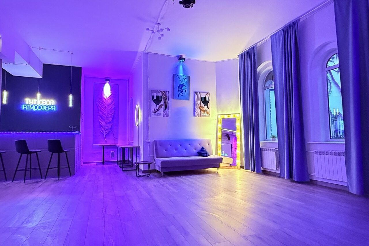 Арендовать Лофт Лайф. Просторное помещение с диваном и зеркалом, украшенное неоновыми вывесками и картинами. Барная стойка с двумя стульями. Большие окна с занавесками, деревянный пол, фиолетовое и жёлтое освещение.