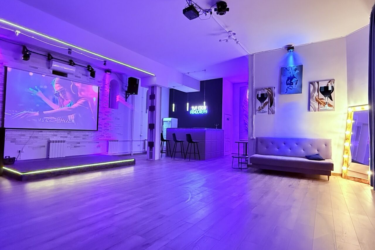Арендовать Лофт Лайф. Просторное помещение с деревянным полом, сцена с экраном, барная стойка с высокими стульями, серая софа, неоновая подсветка, стены с постерами и подсветкой, большое зеркало в раме с лампочками, фиолетовое и синее освещение.