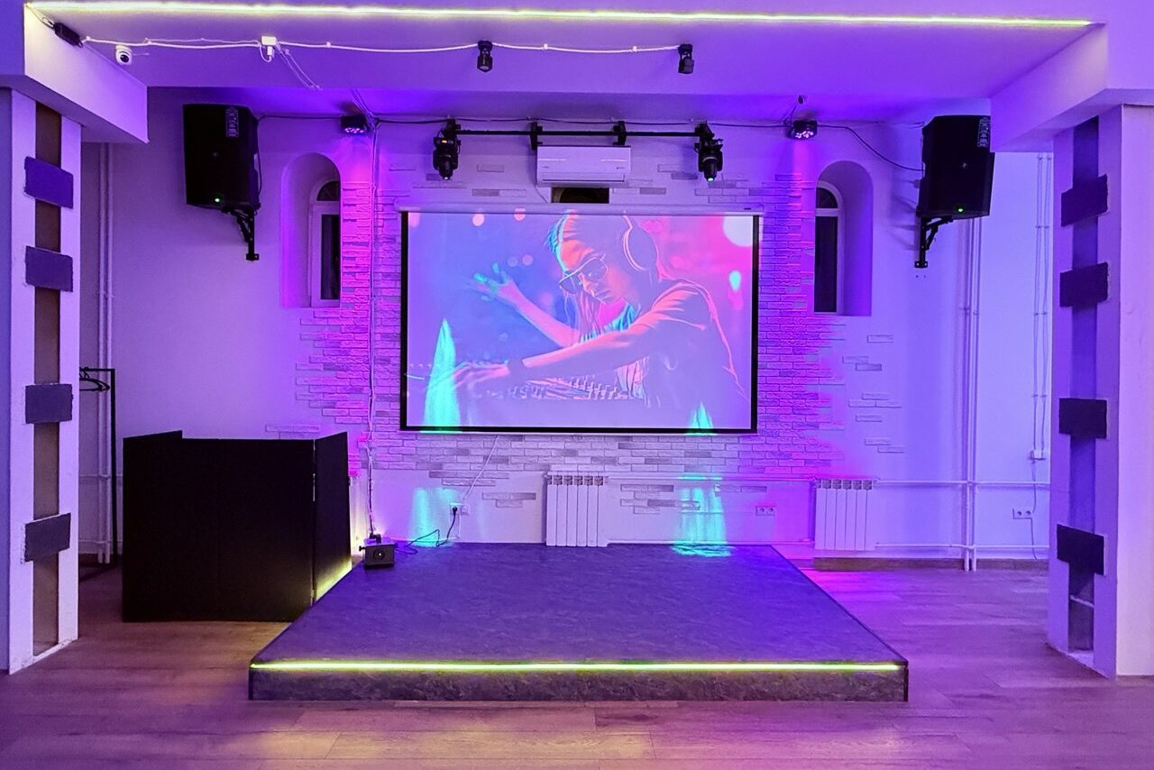 Арендовать Лофт Лайф. Пространство с небольшой сценой, оборудованной экраном и колонками. Потолок с трековыми светильниками, стены белые с отделкой под кирпич, деревянный пол. Сцена подсвечена разноцветным светом, установлен диджейский пульт.
