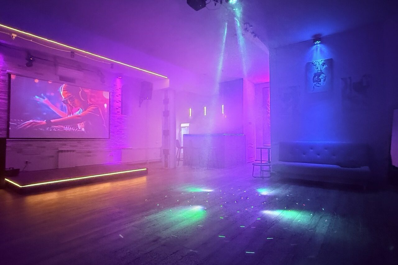 Арендовать Лофт Лайф. Пространство с деревянным полом и сценой, освещенной неоновыми огнями. На стене установлен большой экран с изображением. Справа находится барная стойка и диван. Цветовое освещение меняется от фиолетового до голубого.