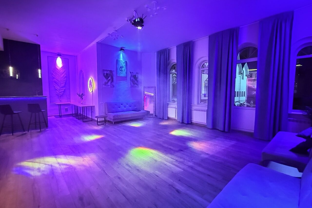 Арендовать Лофт Лайф. Просторное помещение с деревянным полом, рядом барная стойка с двумя стульями. Высокие арочные окна с длинными шторами. Мягкая мебель: несколько диванов. Неоновая подсветка, разнообразное освещение создаёт фиолетовый оттенок. На стенах картины и декор.