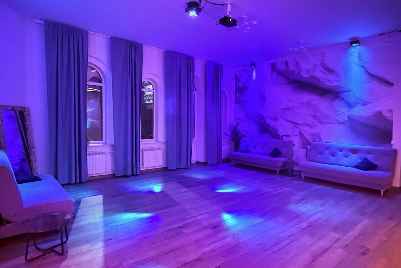 Арендовать Лофт Лайф. Просторное помещение с деревянным полом, двумя диванами у текстурной стены, украшенной эффектом каменной кладки. Три арочных окна с длинными шторами, круглое зеркало, неоновое фиолетовое освещение, потолочные осветительные приборы.