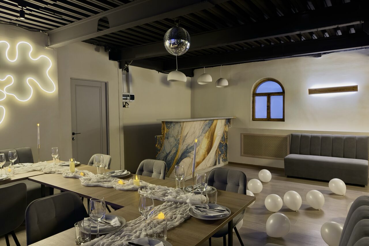 Арендовать Maybe Loft (Мэйби Лофт). Зал Olive (Олив). Современный дизайнерский лофт . Пространство для мероприятий с длинным деревянным столом, сервированным стеклянной посудой и свечами. Мягкие серые стулья, изогнутая серая скамья, стойка с мраморным узором, диско-шар, подвесные светильники, белые шары на полу, декоративная подсветка на стене.