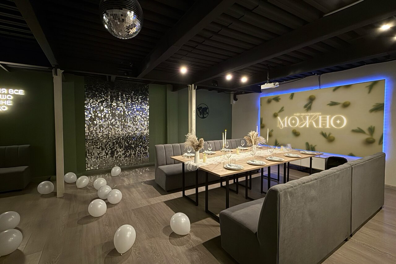 Арендовать Maybe Loft (Мэйби Лофт). Зал Olive (Олив). Современный дизайнерский лофт . Частное помещение с серыми диванами и длинным деревянным столом, украшенным посудой. На полу белые воздушные шары. Неоновая надпись "МОЖНО" на стене с подсветкой. Потолок и пол темные. Зеркальная стена и шар-дискобол под потолком.