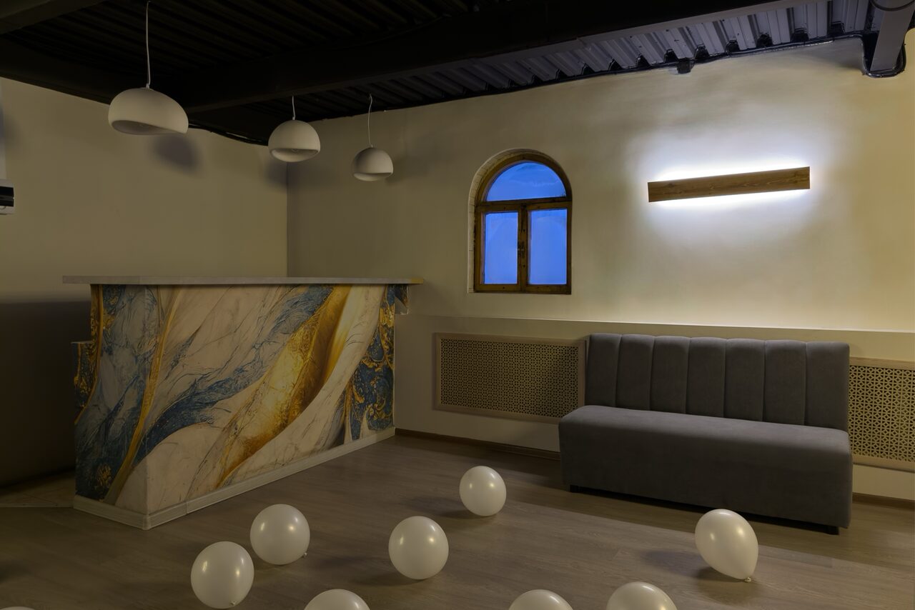 Арендовать Maybe Loft (Мэйби Лофт). Зал Olive (Олив). Современный дизайнерский лофт . Помещение с барной стойкой с абстрактным узором, рядом серая диванная скамья. Потолок декорирован подвесными светильниками. На полу разбросаны белые шары. Стена бежевого цвета с арочным окном и светодиодной панелью.