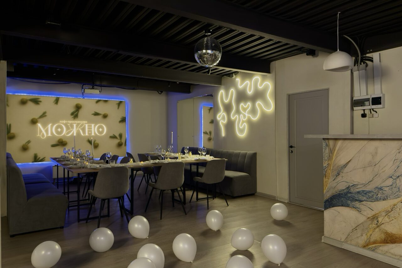 Арендовать Maybe Loft (Мэйби Лофт). Зал Olive (Олив). Современный дизайнерский лофт . Современное пространство с длинным столом и серыми стульями, на столе накрыты приборы и свечи. Стены украшены неоновыми знаками и элементами. Потолок черный, на полу белые шары. Барная стойка с мраморной текстурой и декоративное освещение.
