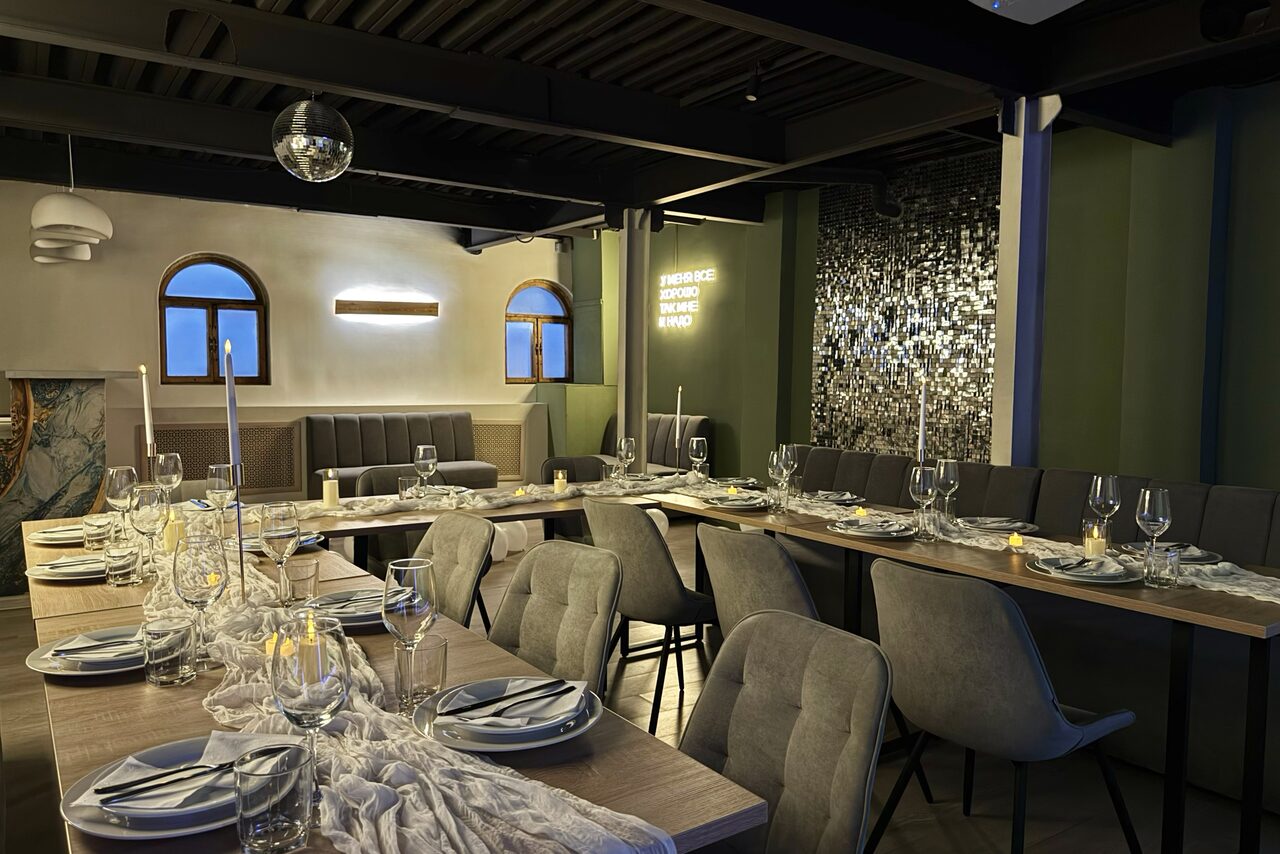 Арендовать Maybe Loft (Мэйби Лофт). Зал Olive (Олив). Современный дизайнерский лофт . Пространство для мероприятий с расставленными столами и стульями, сервированными тарелками, бокалами и свечами. На стене декор из зеркальной плитки и надпись неоном. Светлые и тёмные тона, окна с арочными проёмами, мягкие сиденья.
