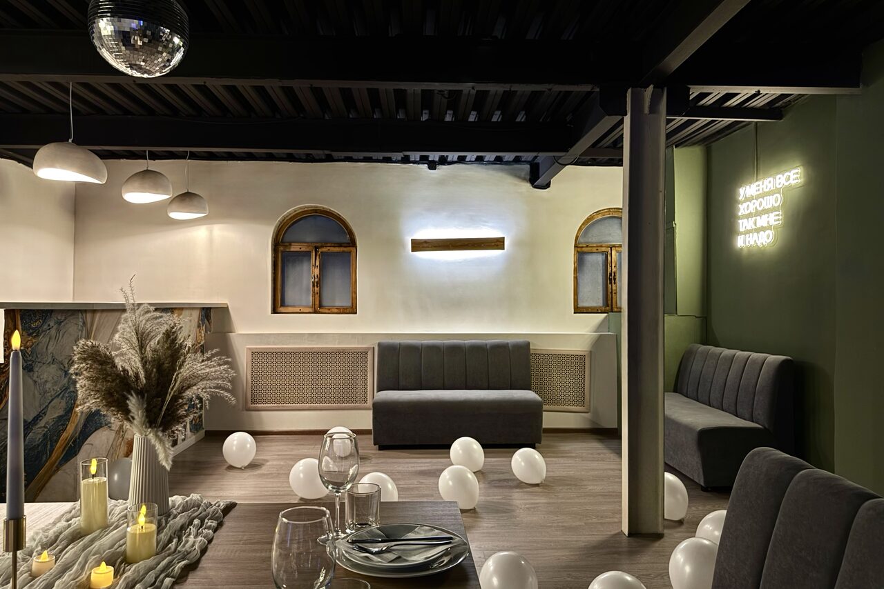 Арендовать Maybe Loft (Мэйби Лофт). Зал Olive (Олив). Современный дизайнерский лофт . Лофт-пространство с деревянным полом и темными потолочными балками, диваны вдоль стен, круглые подвесные светильники, неоновая надпись на стене, декор стола с свечами и сухими растениями, белые шары на полу, два арочных окна.