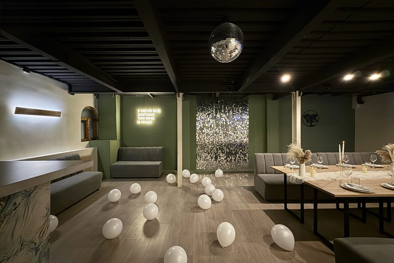 Арендовать Maybe Loft (Мэйби Лофт). Зал Olive (Олив). Современный дизайнерский лофт . Современное пространство с обеденной зоной. Длинный деревянный стол с сервировкой, серые диваны вдоль стен, белые воздушные шары на полу. На стене неоновая надпись, декор из зеркальной плитки. Потолок с черными балками, диско-шар.