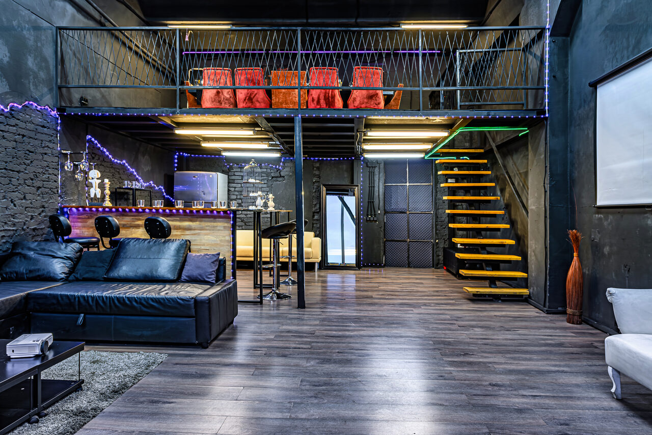 Арендовать Лофт Панорамный. Лофт-пространство с антресолью, черные стены, серый деревянный пол. На первом уровне: черный диван, барная стойка с табуретами, неоновая подсветка, телевизор. Желтая лестница ведет на второй уровень с красными креслами. Мягкое освещение, проектор справа.