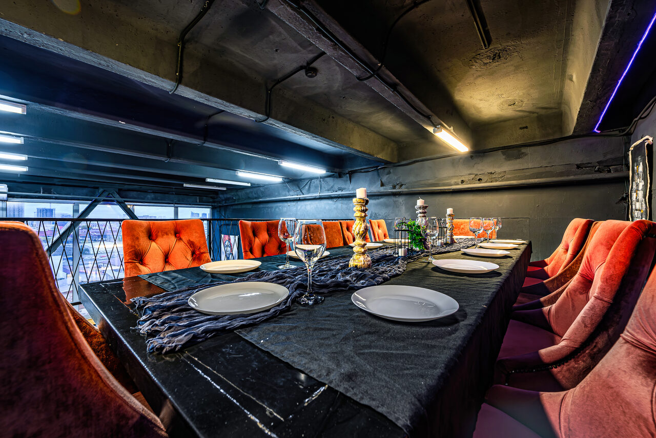 Арендовать Лофт Панорамный. Столовая с длинным черным столом, покрытым темной тканью и сервированным белыми тарелками и бокалами. Вокруг стола - оранжевые мягкие стулья. Потолочные светильники, черный потолок, стены и металлические конструкции. Подсвечники и украшения на столе.