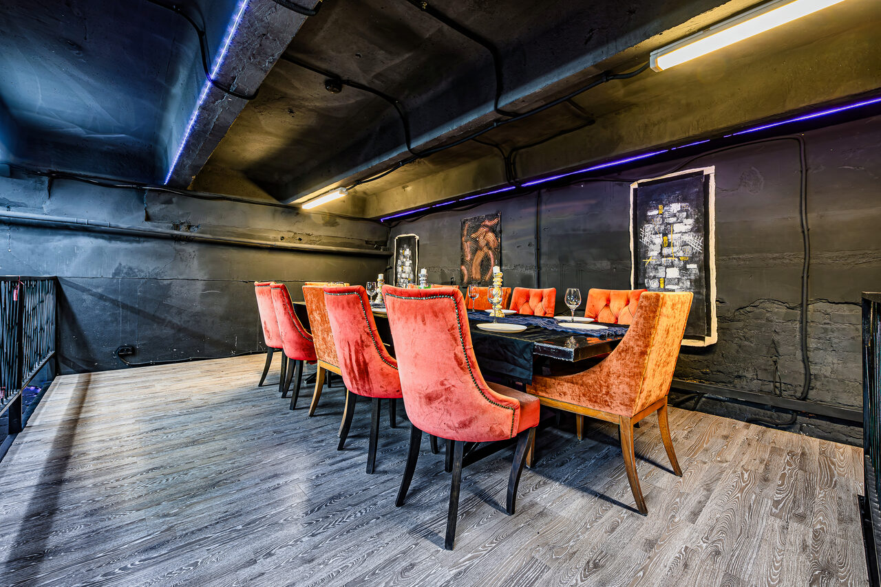 Арендовать Лофт Панорамный. Пространство с черными стенами и потолком, пол серый деревянный. Центральный элемент — темный стол с красными стульями с высокой спинкой. Освещение из длинных светильников на потолке. По стенам висят картины.