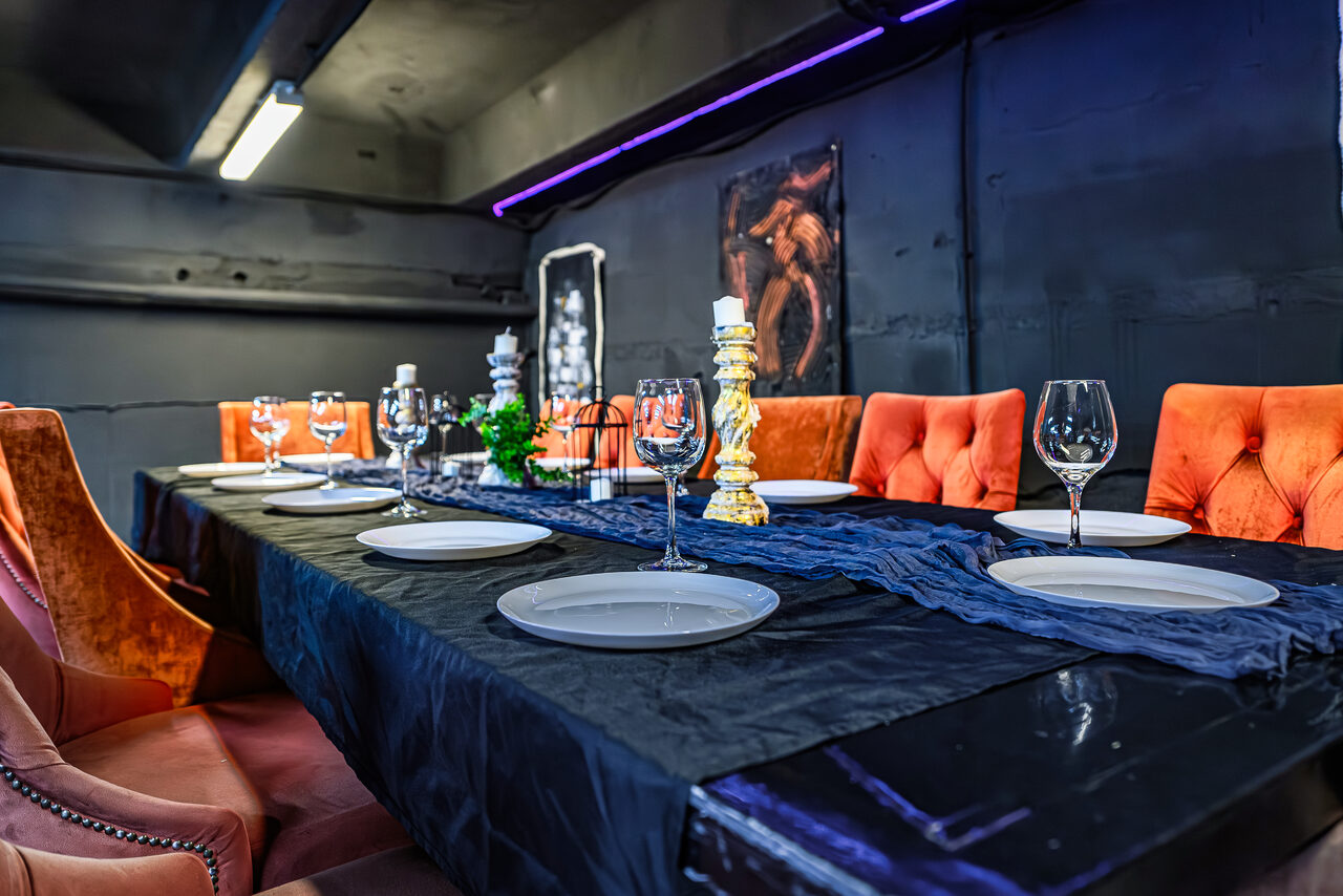 Арендовать Лофт Панорамный. Тёмное помещение с длинным столом, покрытым чёрной скатертью. На столе тарелки, бокалы, высокий подсвечник и декор. Оранжевые и коричневые стулья с мягкой обивкой. Верхний подвесной светильник и фиолетовая подсветка у стены. Стены тёмные.