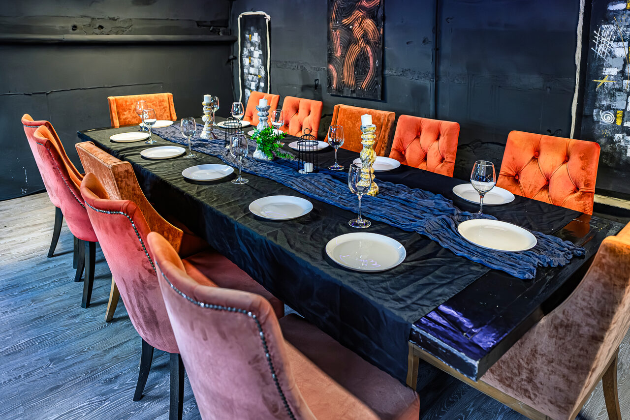 Арендовать Лофт Панорамный. Прямоугольный обеденный стол с черной скатертью, сервированный белыми тарелками и бокалами. Оранжевые и розовые стулья с мягкой обивкой. Темные стены с декором, деревянный пол, центральный элемент — синяя ткань.