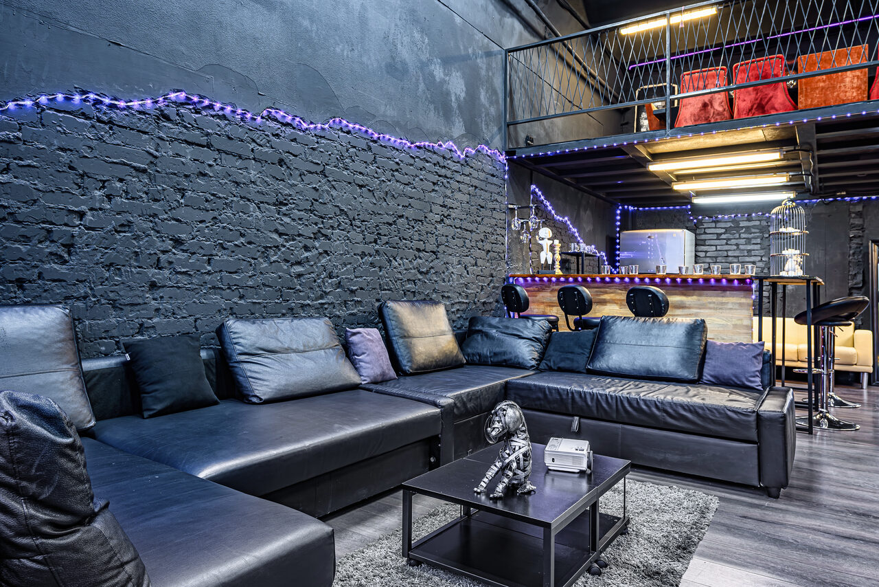 Арендовать Лофт Панорамный. Loft пространство с черными кирпичными стенами, большой угловой кожаный диван, черный журнальный столик, барная зона с высокими стульями и стойкой, неоновая светодиодная подсветка, металлическая лестница на второй уровень, деревянные и металлические элементы интерьера.