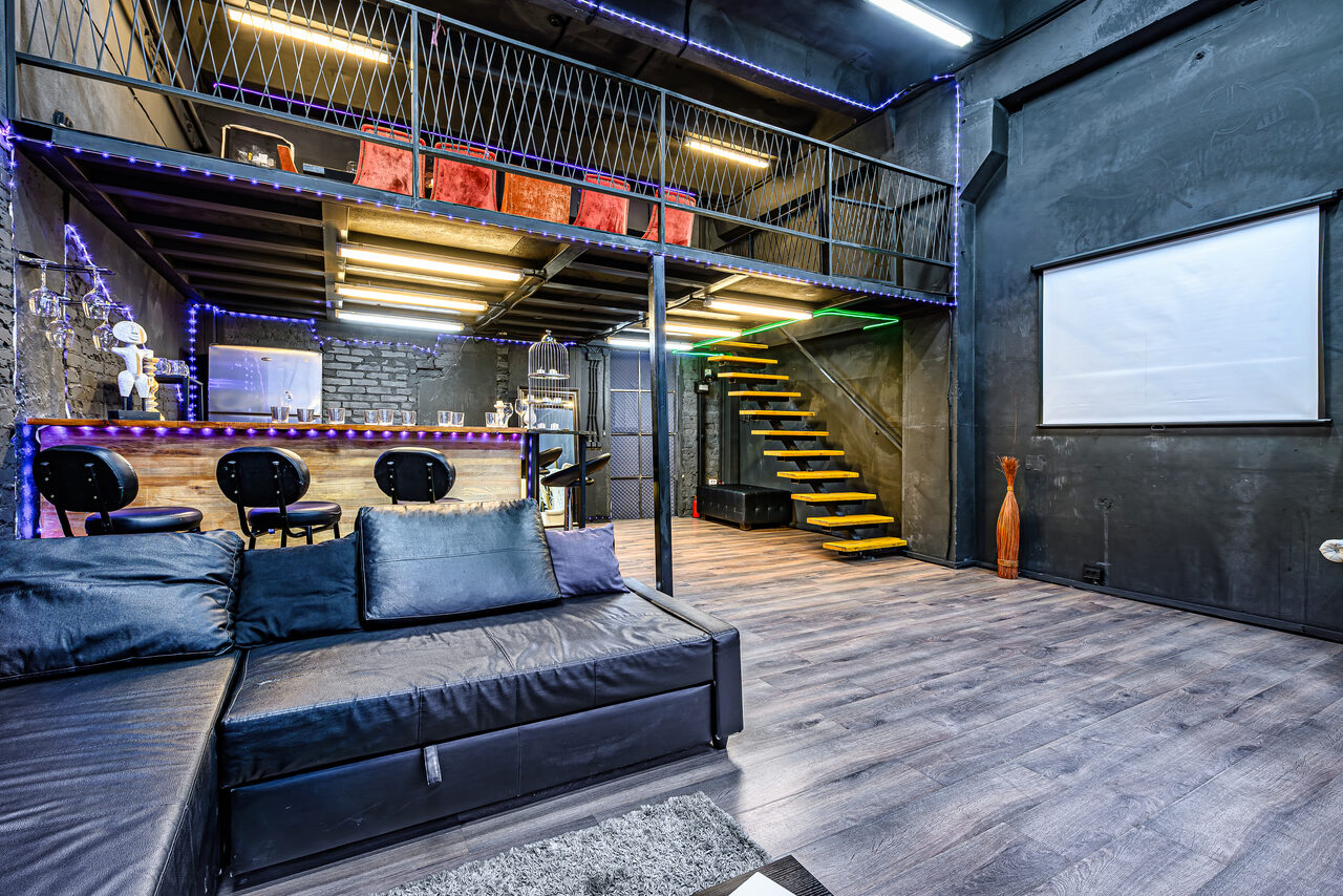 Арендовать Лофт Панорамный. Двухуровневое пространство в стиле лофт с черными стенами и потолками. Внизу черный угловой диван, барная стойка с двумя стульями, флуоресцентное освещение. Наверх ведет лестница с желтыми ступенями. На стене проекторный экран. Пол светлый, деревянный.