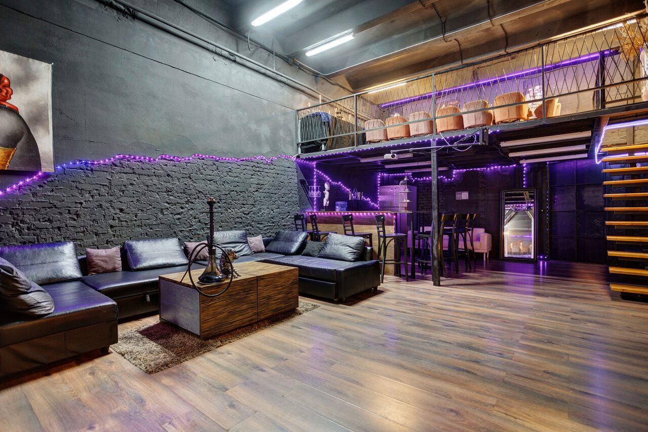 Арендовать Лофт Панорамный. Open industrial-style space with dark gray brick walls, loft area with railing. Black leather sofas, wooden coffee table, and a hookah on a rug. Dining area with high chairs and table. Purple LED lights accentuate the room. Wooden flooring and stairs.