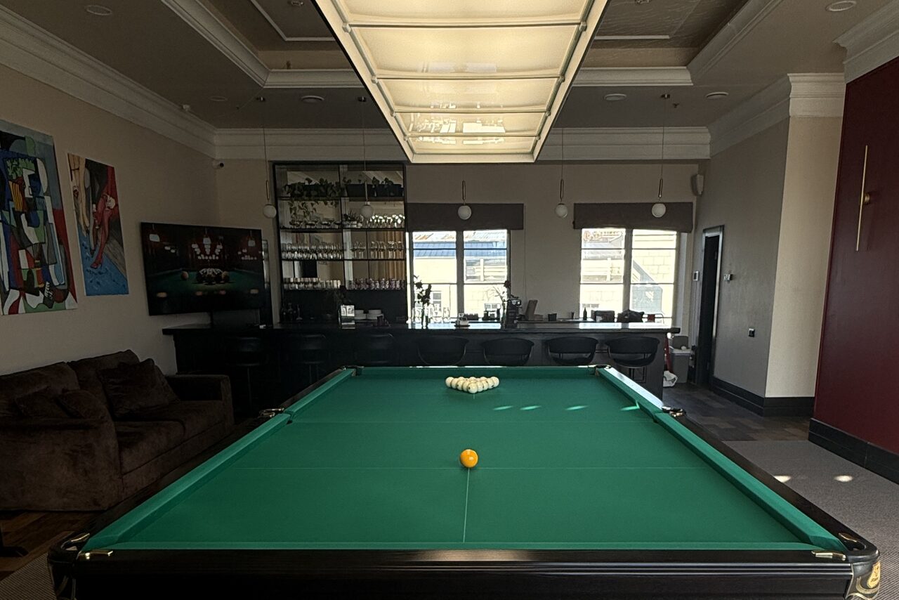 Арендовать Студия Н. Бильярдная комната с зеленым бильярдным столом в центре. На заднем плане барная стойка с высокими стульями и полки с бокалами. На стене висят картины. Освещение: потолочный светильник над столом. Цвета: темные и нейтральные.