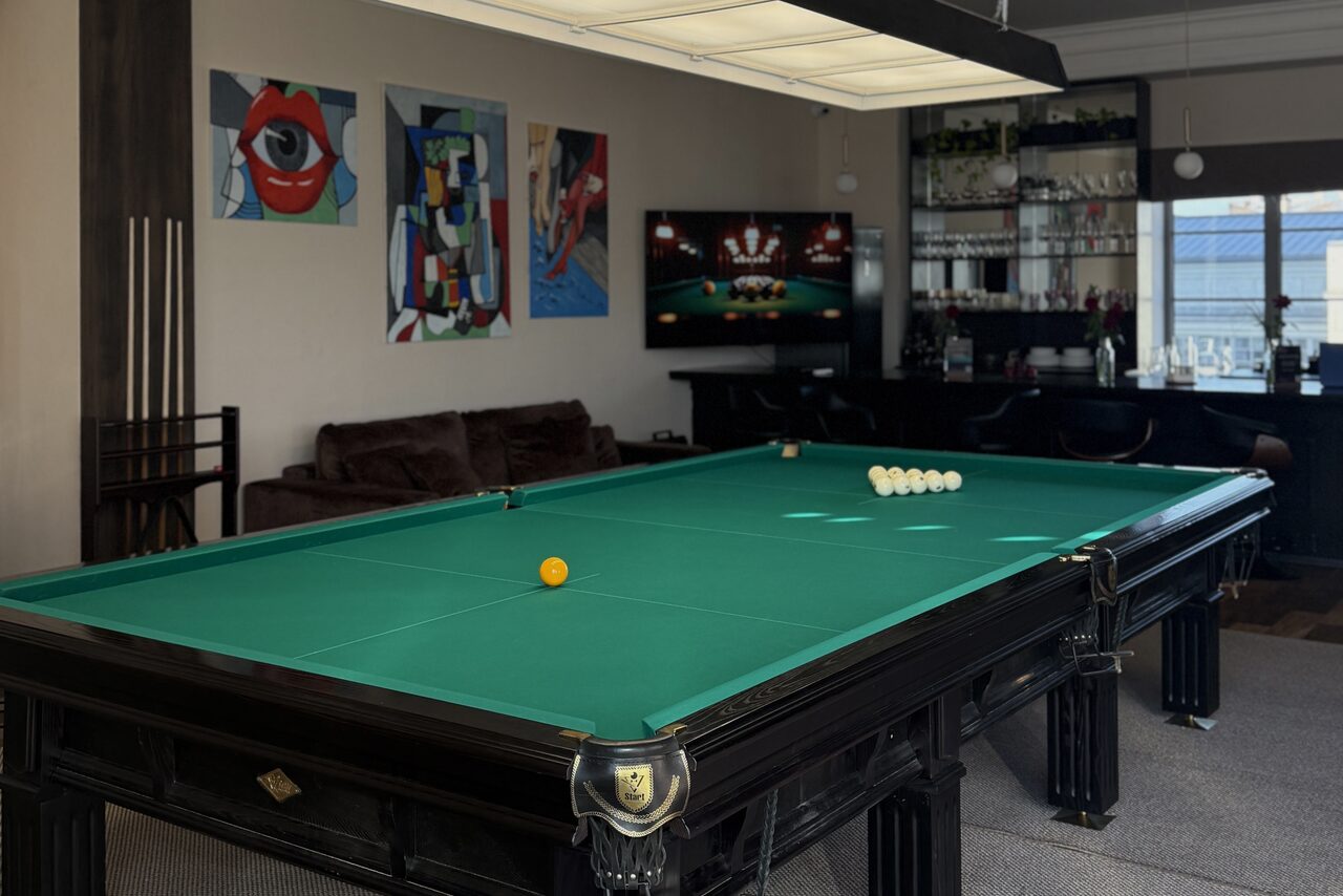 Арендовать Студия Н. Бильярдная с зелёным столом и шарами, рядом диван, на стенах картины. В углу стеллаж с бутылками, бокалом и телевизором. Светлый потолок с подвесным светильником. Холодный свет от окна. Светлые стены, ковровое покрытие пола.