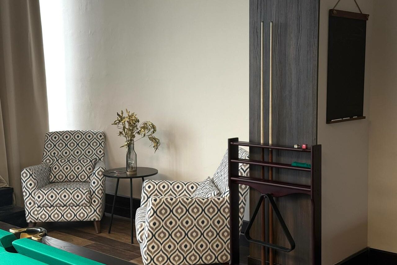 Арендовать Студия Н. Комната с двумя креслами с геометрическим узором, круглым столиком с вазой, стойкой для бильярдных киев, черной колонкой и небольшим настенным меловым табло. Стены светлые, пол деревянный темного оттенка.