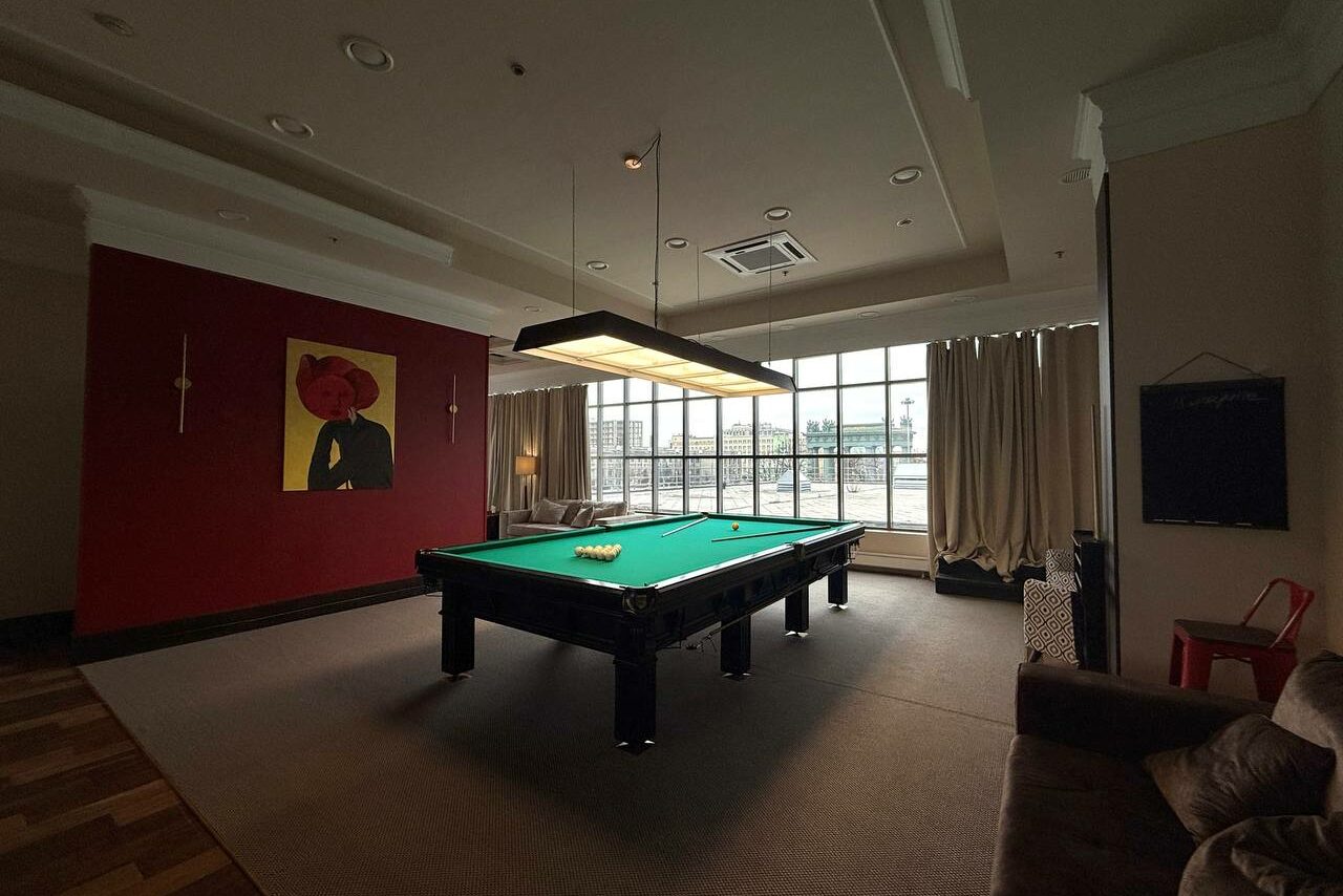 Арендовать Студия Н. Просторная комната с бильярдным столом в центре, красная стена с картинами, большая панорамная витражная окно с занавесками, мягкий диван и кресло, подвесной светильник над столом, паркетный пол.