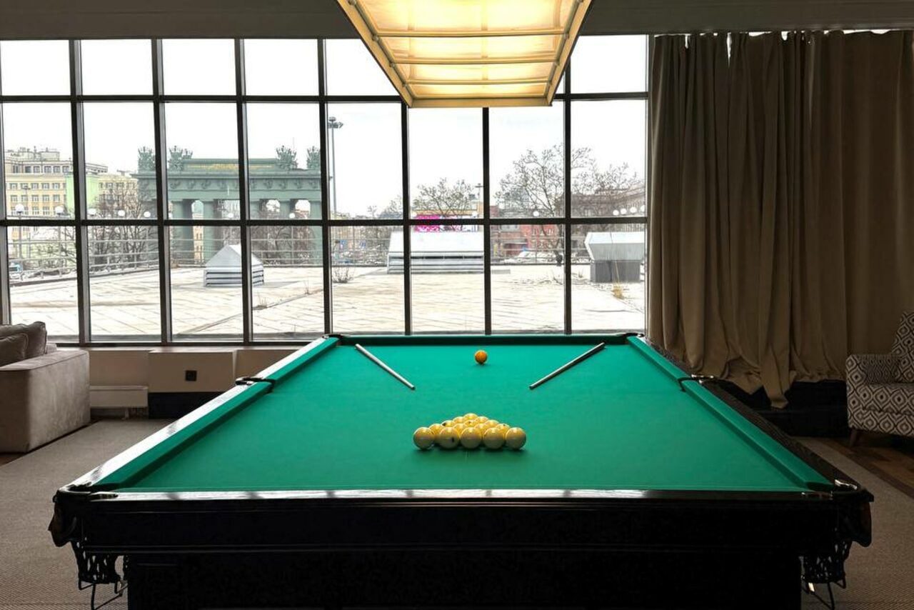 Арендовать Студия Н. Бильярдная комната с большим столом, зеленым сукном, двумя киями и набором желтых шаров. За столом панорамные окна с видом на улицу. Слева диван, справа кресло с узором. Потолочный светильник, бежевые шторы.