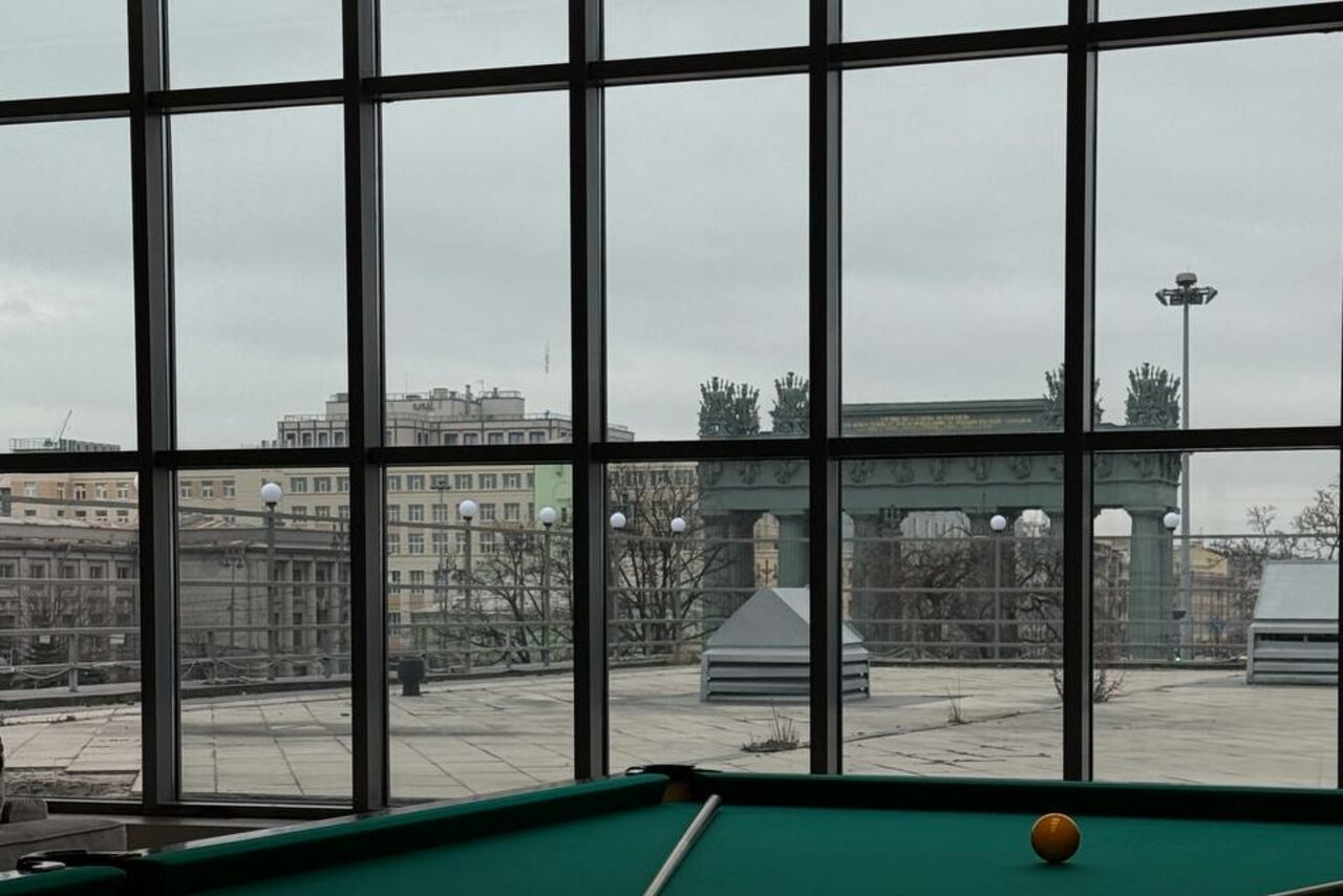 Арендовать Студия Н. Пространство с большими панорамными окнами, за которыми видна терраса и городская архитектура. Внизу части окна расположен стол для бильярда с зеленым сукном и шаром. Потолочный светильник не включен. Цвета: серый, зеленый.