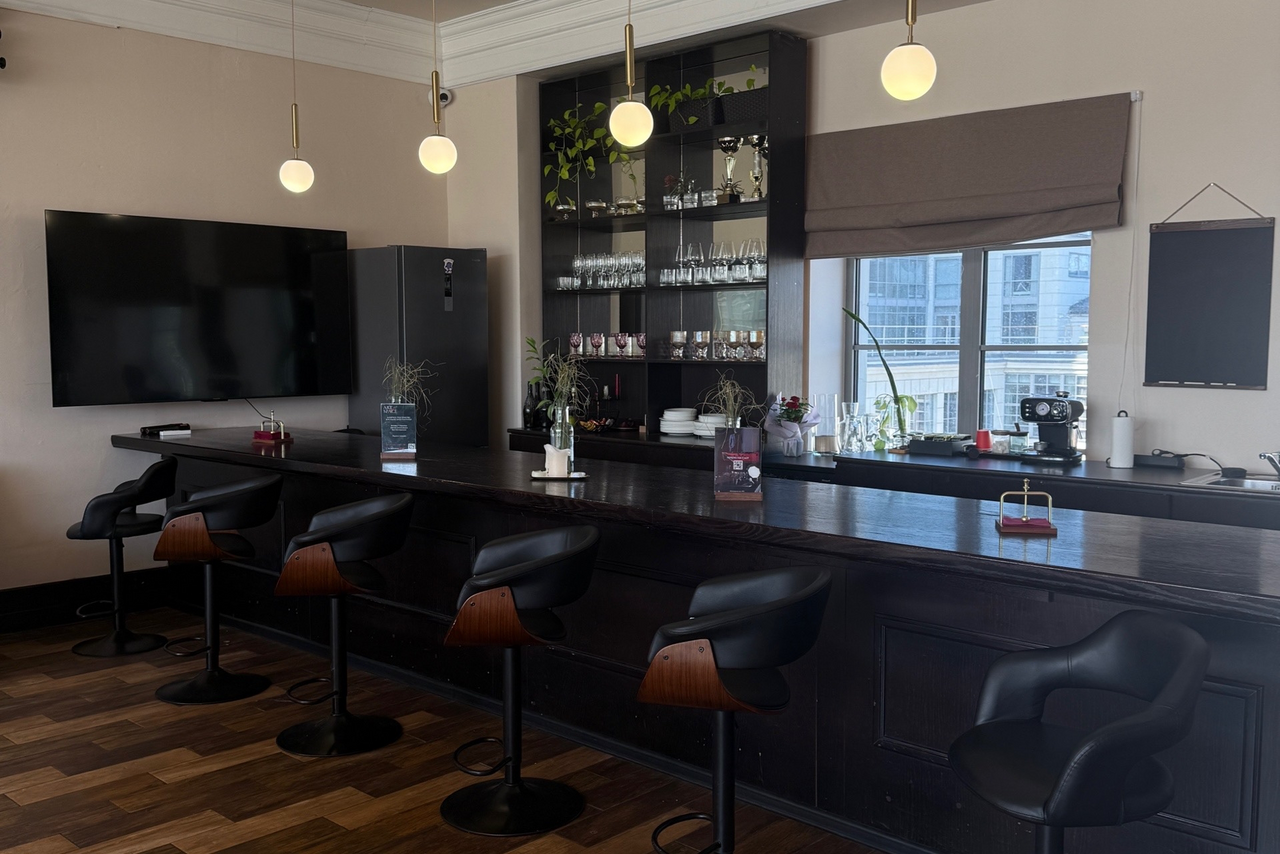 Арендовать Студия Н. Барная стойка с черными стульями и темной деревянной отделкой, высокие шкафы с посудой и декором, телевизор, холодильник, подвесные светильники, окно с римской шторой, на стойке растения, таблички и диспенсеры для салфеток.