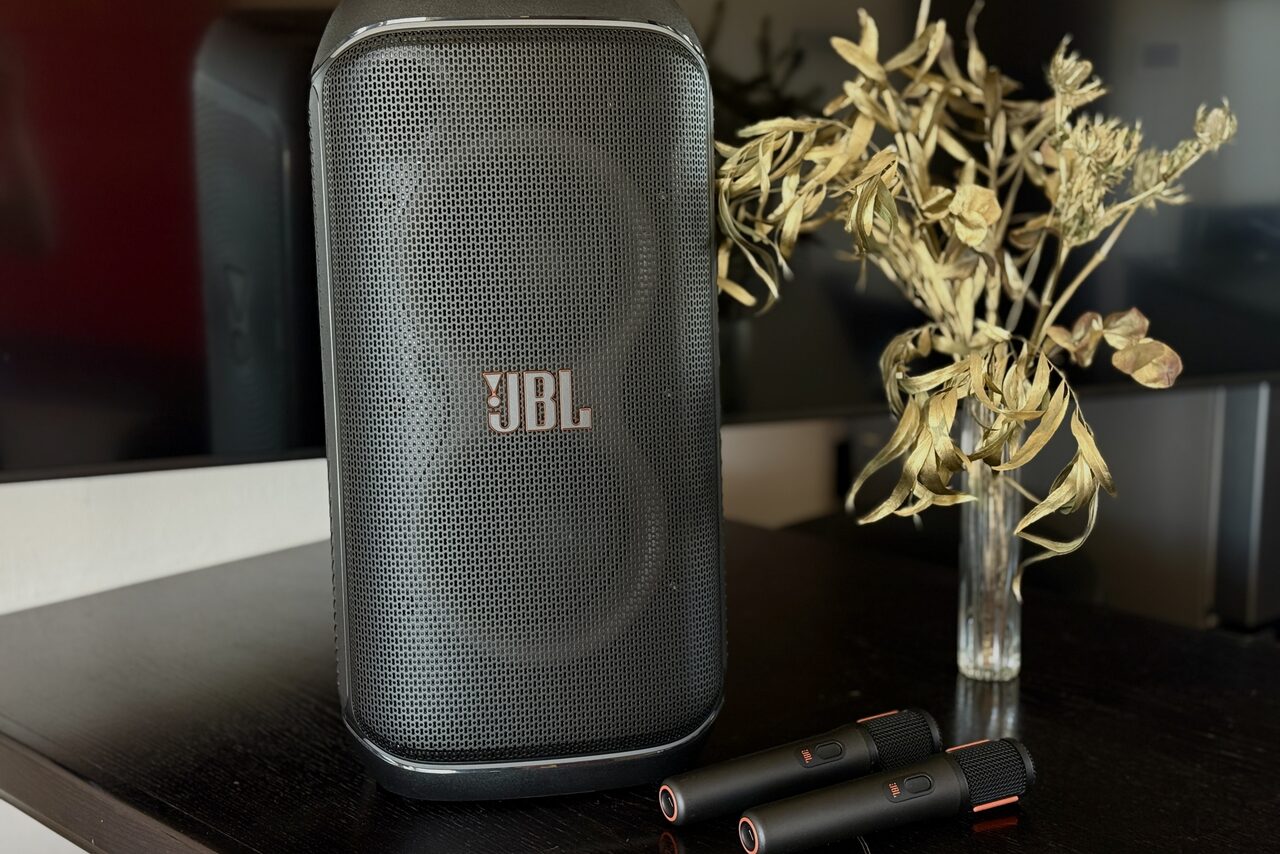 Арендовать Студия Н. Черная аудиоколонка на темном столе, рядом с ней два микрофона и ваза с сухими растениями. На заднем фоне видна часть телевизора.