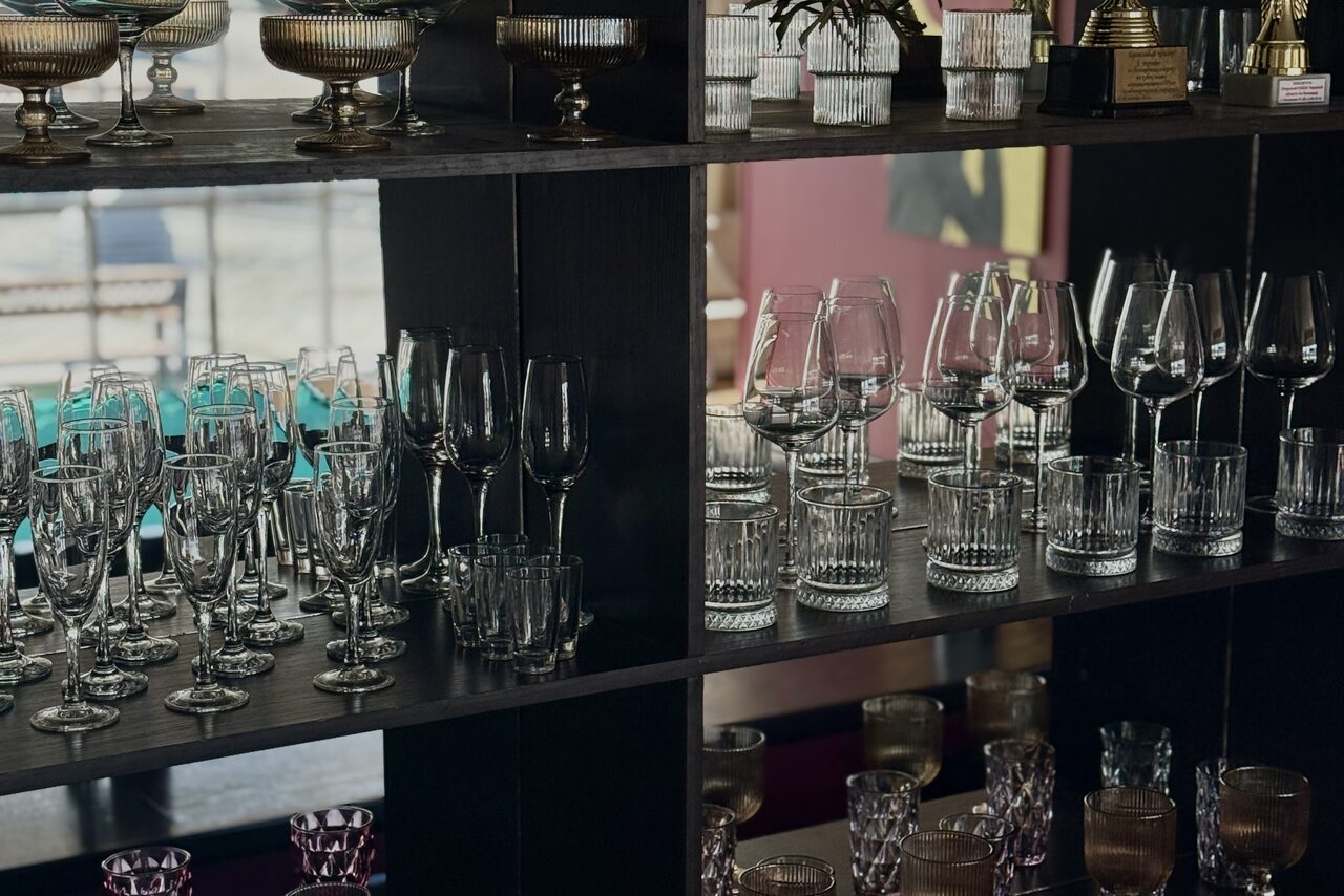 Арендовать Студия Н. Полка с разнообразными бокалами и стаканами. Черная деревянная конструкция, разделённая на секции. На заднем плане виден размытый интерьер с розовыми и бирюзовыми элементами. Естественное освещение через окно слева.