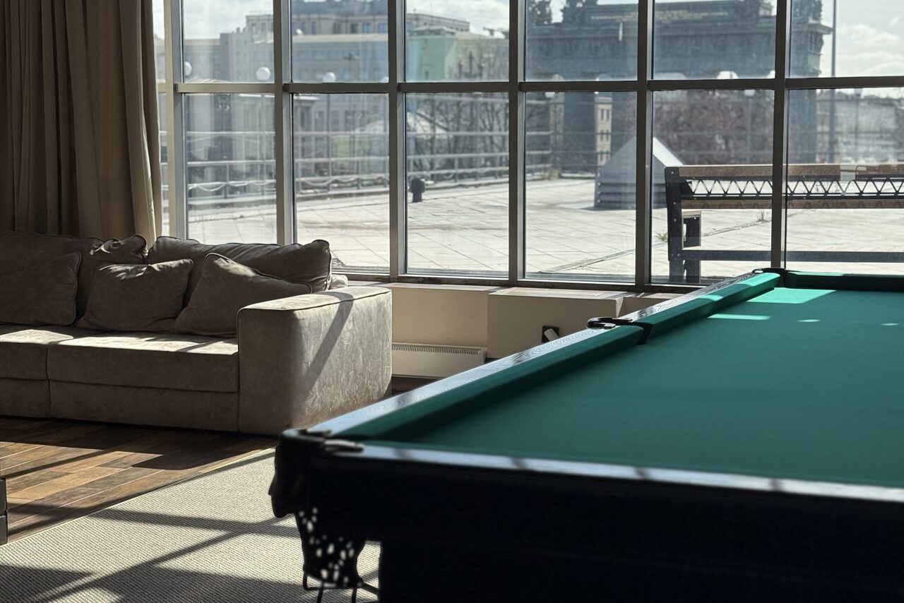 Арендовать Студия Н. Просторное помещение с большими окнами и видом на город. В центре зеленый бильярдный стол. Справа светлая мягкая мебель. Напольное покрытие сочетает ковровую и паркетную зоны. Естественное освещение, светлые занавески.